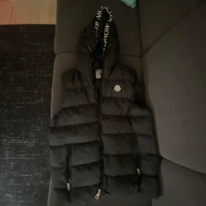 Svart Moncler Väst - Snygg svart dunväst från Moncler med stor huva och logga på bröstet. Västen har dragkedja framtill, två fickor med dragkedja och Moncler-tryck på huvkanten. Perfekt för lager-på-lager och streetstyle.
