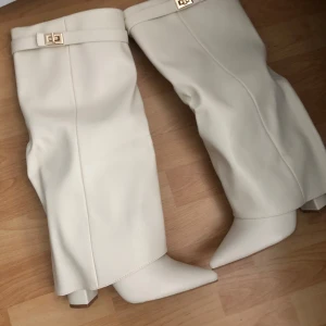 Vita stövlar med gulddetalj - Snygga vita stövlar med spetsig tå och låg klack. Tillverkade i skinnliknande material och dekorerade med en elegant guldfärgad spänndetalj upptill. Perfekta för dig som vill sticka ut med en clean och trendig look.