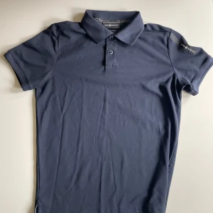 Mörkblå pikétröja Sail Racing - Snygg mörkblå pikétröja från Sail Racing med klassisk krage och två knappar framtill. Tröjan har korta ärmar och diskret logga på ärmen. Tillverkad i mjuk bomull som känns skön mot huden. Perfekt för en clean och sportig stil. Säljer för 300kr