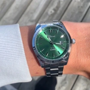 Grön klocka med stållänk från Doventi - Snygg klocka från Doventi med grön urtavla och datumvisning. Klockan har en rund boett i silverfärgat stål och matchande länkarmband. Stilren design som sticker ut med sin gröna färg och metalliska finish. Perfekt accessoar för dig som vill ha något unikt på handleden.