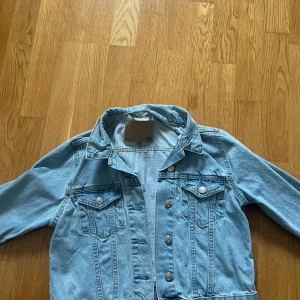 Ljusblå jeansjacka XS med knappar - Säljer en klassisk ljusblå jeansjacka från 157 i storlek XS. Jackan har silverfärgade knappar framtill, två bröstfickor med lock och knapp samt en snygg krage. Perfekt att slänga över en hoodie eller t-shirt för en chill look.