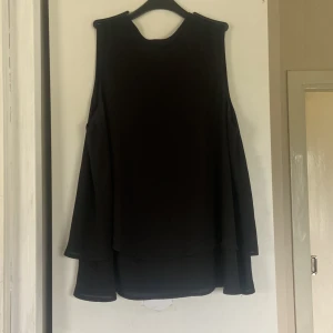 Svart blus med capeärm från Zizzi - Svart blus från Zizzi med unika capeärmar och rund halsringning. Blusen har en lös passform och är tillverkad i ett lätt, flowy tyg som ger ett snyggt fall. Perfekt för dig som gillar stilrena och coola plagg med en twist.