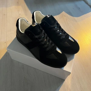 Maison Margiela svarta sneakers i skinn - Maison Margiela sneakers i svart skinn och mocka med klassisk låg siluett. Skorna har svarta snören, vit insida och diskreta detaljer i svart. Snygg och stilren design med rund tå och platt sula. Kommer med originalkartong och dustbag.