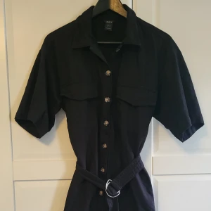 Svart skjortklänning från Lindex - Svart kort skjortklänning från Lindex med knappar hela vägen framtill, två bröstfickor och tillhörande bälte i midjan. Klänningen har korta ärmar och klassisk krage, perfekt för en stilren look. Använd kanske två gånger, passar en storlek 36-38. Öppen för prisförslag, hör av dig med frågor 🌸