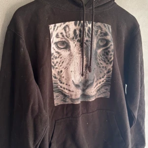 Svart hoodie med leopardtryck - Svart hoodie med stor leopardbild framtill. Tröjan har huva med snörning och en klassisk ficka ifram.