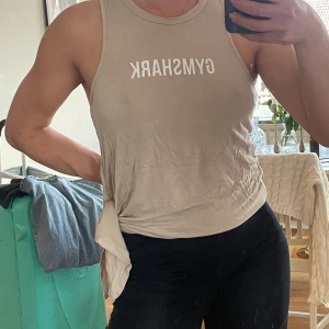 Beige linnetopp från Gymshark - Säljer en snygg beige linnetopp från Gymshark med vitt tryck framtill. Toppen är ärmlös och har en rund halsringning. Perfekt för träning eller chill dagar. Materialet är mjukt och lätt, vilket gör den superbekväm att bära.