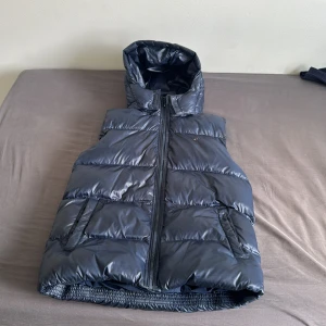 Mörkblå dunväst Tommy Hilfiger - Snygg mörkblå dunväst från Tommy Hilfiger med huva och dragkedja. Västen har stora quiltade partier, två fickor fram och klassisk Hilfiger-logga i rött och vitt på ryggen. Materialet är glansigt och känns riktigt sportigt och modernt. 