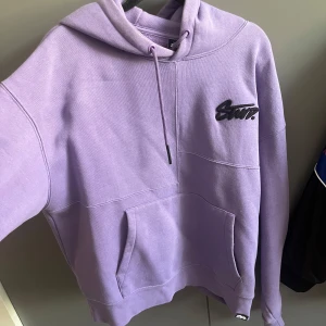 Ljuslila hoodie från STWD med huva - Säljer en ljuslila hoodie från STWD med broderad logga på bröstet och tryck på huvan. Tröjan har stor ficka framtill, snörning i huvan och en praktisk dragkedja i sidan. Perfekt för dig som gillar streetwear och vill sticka ut lite extra. Sista bilden visar färgfläck.