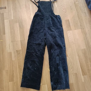 Svart sammets byxdress med vida ben - Säljer en svart byxdress i mjuk sammet med raka, vida ben och justerbara axelband. Byxdressen har fickor både fram och bak, perfekt för en chill och avslappnad stil. Passar dig som gillar retrovibbar och vill ha något bekvämt men ändå snyggt.