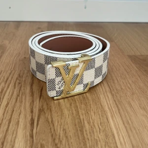 Louis Vuitton rutig skärp med LV-spänne - Snyggt Louis Vuitton-bälte i vitt och grått med rutigt mönster och ikoniskt LV-spänne i guld. Bältet är gjort i skinn och har klassisk design med tydlig logga. Perfekt accessoar för att lyfta din outfit med lyxig känsla.