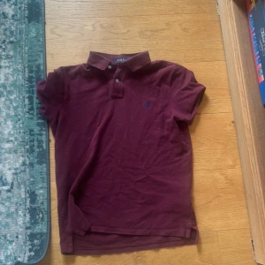 Vinröd pikétröja från Polo Ralph Lauren - Säljer en vinröd pikétröja från Polo Ralph Lauren med klassisk krage och två knappar framtill. Tröjan har korta ärmar och den ikoniska blå loggan broderad på bröstet. Modellen är custom slim fit och tillverkad i mjuk bomull. Perfekt för en clean och stilren look.