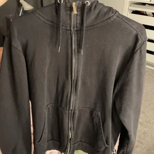 Svart hoodie med dragkedja och huva - En svart hoodie med dragkedja framtill och huva med snörning. Tröjan har långa ärmar och två stora fickor framtill. Perfekt för chill dagar eller när du vill ha en enkel och stilren look.