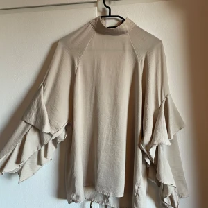 Beige blus med volangärmar H&M, stl 34 -  beige blus från H&M med höghalsad krage och knappar i nacken. Blusen har långa, vida ärmar med flera lager volanger storlek 34 