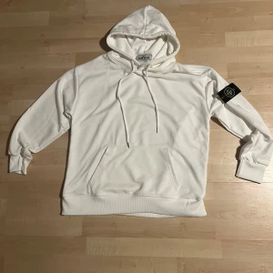 Vit hoodie från Stone Island - Snygg vit hoodie från Stone Island med klassisk logga på vänster ärm. Tröjan har huva med snörning, känguruficka framtill och ribbade muddar. Perfekt för dig som gillar clean och stilren streetwear. Oanvänd, pris kan diskuteras 