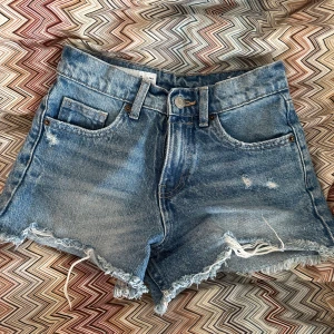Blå jeansshorts med fransig kant från Zara - Snygga blå jeansshorts från Zara med fransig rå kant och slitna detaljer framtill. Klassisk femficksmodell med bälteshällor och knappstängning. Perfekta för en chill sommarlook.