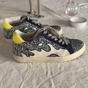 Glittriga sneakers från Bibi Lou - Snygga sneakers från Bibi Lou med blått glitter över hela skon, beige mockadetaljer och blåa partier i mocka. Hälkappan är i en poppig gul färg. Skorna har snörning och en platt vit sula. Perfekta för dig som vill sticka ut med coola detaljer.