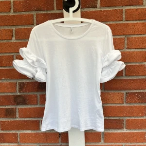 Vit volangtopp från ONLY - Superfin vit topp från ONLY med korta ärmar och snygga volangdetaljer på ärmarna. Rund halsringning och mjukt material som känns skönt mot huden. Perfekt för dig som vill ha en enkel men ändå unik look.