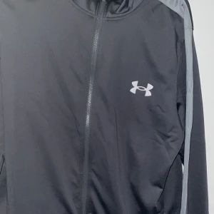 Grå kofta från Under Armour - Grå kofta från Under Armour med gråa detaljer längs ärmarna och dragkedja framtill. Jackan har en sportig look med logga på bröstet och är tillverkad i ett lätt syntetmaterial. Perfekt för dig som gillar att hålla stilen på gymmet eller ute.