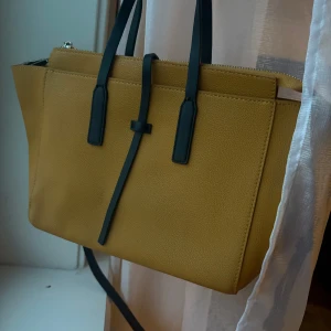 Gul handväska från Zara - Stilren handväska från Zara i senapsgult skinnmaterial med svarta handtag och axelrem. Väskan har en klassisk fyrkantig form, dragkedja upptill och minimalistisk design. Rymmer väldigt mycket.