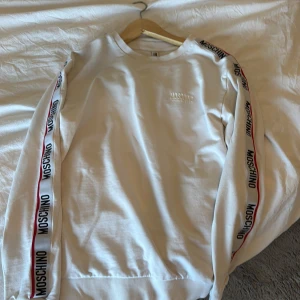 Vit Moschino sweatshirt med logoband - Snygg vit sweatshirt från Moschino med logoband längs båda ärmarna och diskret broderad logga på bröstet. Tröjan har rund hals, ribbade muddar och är tillverkad i mjuk bomull. Perfekt för en clean och trendig look.
