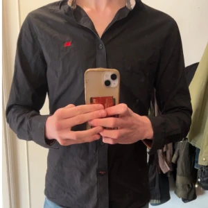 Svart Burberry skjorta med rutiga detaljer - Snygg svart trendig skjorta från Burberry med klassiskt rutigt mönster på insidan av kragen och ärmsluten. Perfekt för en stilren look. Storlek M använd väl. Styrk, tvätta den och den ser ut som ny! 