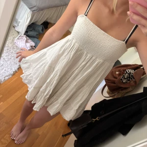 Vit kort sommarklänning från Zara - Supersöt vit kort klänning från Zara med smock upptill och tunna axelband. Klänningen har en flowy, volangig kjol och är perfekt för varma dagar. Lätt och luftig i bomull, passar dig som gillar en avslappnad och feminin stil. Passar som s