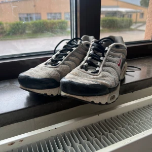 Nike Air Max Plus Tn sneakers grå/vit - Nike Air Max Plus Tn sneakers i grått och vitt med svarta och röda detaljer. Skorna har mesh-överdel, svarta snören och synlig Air Max-dämpning i sulan. Ikonisk Tn Air-logga bak och vågiga linjer på sidorna ger en sportig vibe.