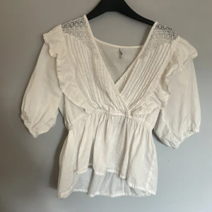 Vit blus med volang från JDY - Supersöt vit blus från JDY med v-ringning, volangdetaljer och spets vid axlarna. Blusen har korta puffärmar och smock i midjan för en snygg passform. Perfekt för dig som gillar romantisk och bohemisk stil.