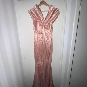 Lång glansig champagnefärgad klänning - Säljer en elegant långklänning i glansigt champagnefärgat tyg. Klänningen har en omlottliknande drapering upptill och är ärmlös med breda axelband. Materialet ger en lyxig känsla och passar perfekt för speciella tillfällen.