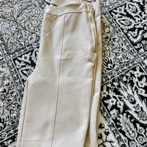 Beiga skinnleggings med dragkedja - Snygga beiga leggings i skinnimitation med dragkedja vid midjan och slits nertill på benen. Tight passform och stilrena sömmar längs framsidan. Perfekta för en trendig och edgy look. Helt nya aldrig använda ,stl S 