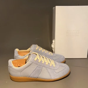 Maison Margiela ljusgrå sneakers - Maison Margiela sneakers i ljusgrått skinn och mocka med klassisk gummisula och vita snören. Snygg minimalistisk design med diskreta paneler och retrovibbar. Perfekta för dig som gillar stilrena och exklusiva sneakers.