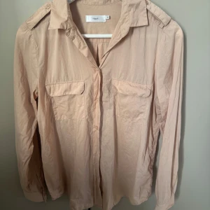 Beige skjorta från Filippa K smal - Stilren beige skjorta från Filippa K med klassisk krage, två bröstfickor med lock och knäppning framtill. Skjortan har långa ärmar och axelklaffar för en cool touch. Perfekt att styla med jeans eller kjol.