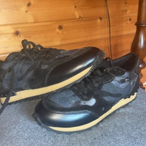 Valentino Garavani Rockrunner sneakers - Svarta Valentino Garavani Rockrunner sneakers med cool camouflagemönstrad ovandel i mocka och skinn, beige sula och ikoniska nitar på hälen. Snygg och edgy design med snörning och rund tå. Perfekta för dig som vill sticka ut med lyxiga detaljer.