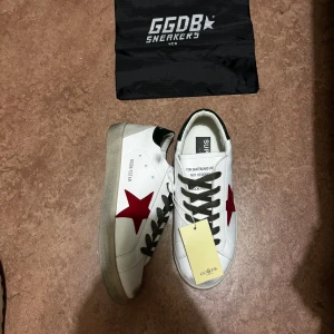 Golden Goose sneakers - Golden Goose sneakers i vitt skinn med ikonisk röd stjärna på sidan, gröna detaljer på hälen och svarta snören. Snygg kontrast mellan den vita ovandelen och den gråtonade sulan. Klassisk rund tå och platt sula, perfekt för en cool streetstil.