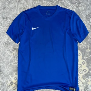 Blå Nike Dri-Fit t-shirt - Sportig blå t-shirt från Nike med Dri-Fit-teknologi. Klassisk passform med rund hals och korta ärmar. Swoosh-logga i vitt på bröstet. Perfekt för träning eller när du vill ha en clean och bekväm stil.