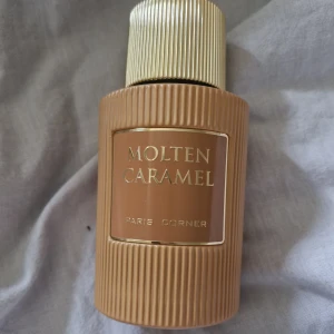 Molten Caramel Paris Corner parfym - Molten Caramel från Paris Corner är en lyxig eau de parfum i en snygg beige och guldig flaska med räfflad design. Flaskan har en rund form och detaljer i guld, vilket ger en exklusiv känsla. Doften är inspirerad av karamell och passar dig som gillar söta och varma toner. Testsprutad 