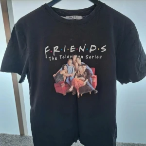 Svart Oversized Friends t-shirt från NA-KD - Svart t-shirt från NA-KD med stort Friends-tryck framtill och avsnittslista på ryggen. Klassisk rund hals, korta ärmar och mjukt bomullsmaterial. Perfekt för dig som älskar serien och vill ha en statement tee med ikoniskt motiv.