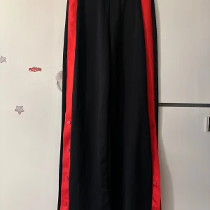 Svarta wide byxor med röda knappar - Säljer ett par svarta wide byxor med röda sidor och tryckknappar längs hela benet. Byxorna har en sportig vibe och är riktigt sköna med lös passform. Perfekta för dig som gillar streetstyle och vill sticka ut lite extra.