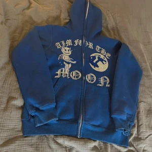 Blå hoodie Aim For The Moon - Blå hoodie med hel dragkedja och huva från Aim For The Moon. Framsidan har ett stort tryck med texten 'AIM FOR THE MOON' och en tecknad figur samt måne i glittriga stenar. Mjuk insida och ribbade muddar. Perfekt för dig som gillar streetwear vibes. Lite nopping