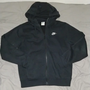 Nike Zip hoodie - Svart Zip hoodie från Nike med vit logga på bröstet. Tröjan har huva med snörning, hel dragkedja framtill och två fickor. Tillverkad i mjuk bomullsblandning. Klassisk och enkel design som funkar året runt. Endast använd nån gång inga spår av använding.