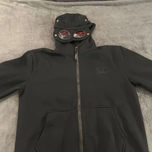 Svart gogglejacka från C.P. Company - Svart jacka från C.P. Company med ikoniska goggle-detaljer i huvan. Jackan har dragkedja framtill, två fickor och broderad logga på bröstet. Tillverkad i mjukt material och har en clean, modern look som sticker ut.