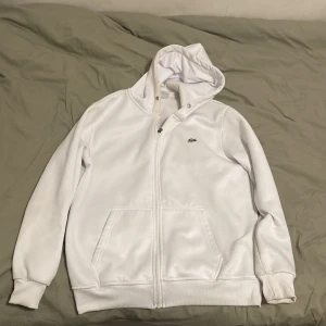 Vit hoodie från Lacoste med dragkedja - Snygg vit hoodie från Lacoste med klassisk krokodillogga på bröstet. Tröjan har huva, dragkedja framtill och två stora fickor. Perfekt för dig som gillar stilren och sportig look. Materialet känns mjukt och skönt mot huden.