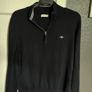 GANT 1/4 Zip - Svart stickad tröja från Gant med halv dragkedja och ribbad krage med vit rand. Broderad Gant-logga på bröstet. Långärmad och klassisk passform, perfekt för en stilren look.