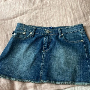 Jeanskjol från Victoria Beckham med brodyr - Snygg blå jeanskjol från Victoria Beckham med slitna kanter och broderade kronor på bakfickorna. Klassisk femficksmodell, bälteshällor och knappstängning. Midjemått: 38cm längd: 31cm
