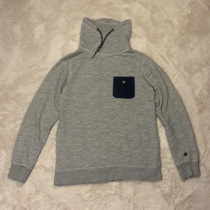 Grå Esprit tröja med hög krage - Snygg grå tröja från Esprit med hög krage och snörning. Tröjan har lång ärm och en mörkblå bröstficka med knapp. Perfekt för kyliga dagar och enkel att matcha med jeans eller joggers.