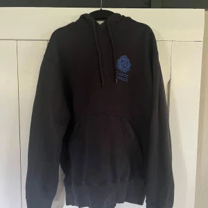 Svart hoodie från Cleptomanicx M - Svart hoodie från Cleptomanicx i storlek medium med blått tryck av händer både fram och bak. Klassisk känguruficka, snörning i huvan och mjukt material. Perfekt för dig som gillar streetwear och vill ha en chill look.