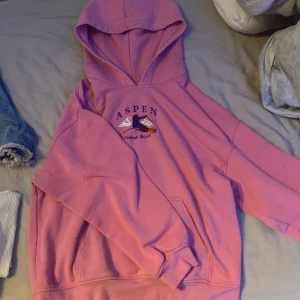 Rosa Aspen hoodie med broderi - Rosa hoodie med broderad skidåkare och texten 'Aspen Colorado, U.S.A.' på bröstet. Tröjan har huva, magficka och långa ärmar. Perfekt för dig som gillar en chill och sportig stil.