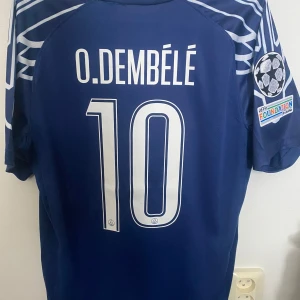 PSG Dembele matchtröja blå - Snygg blå PSG fotbollströja med O. Dembélé och nummer 10 på ryggen. Tröjan har vita detaljer, Qatar Airways-tryck på bröstet, Champions League-märke på ärmen och coola grafiska mönster på axlarna. Tillverkad i lätt och ventilerande material.
