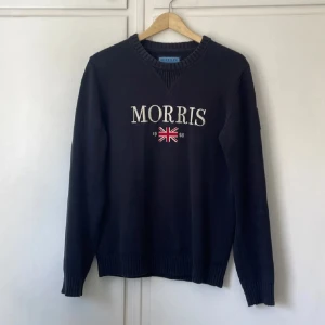 Mörkblå stickad tröja från Morris Co - Snygg mörkblå stickad tröja från Morris Co med broderad logga och brittisk flagga framtill. Tröjan har ribbade muddar, rund hals och en patch på ärmen. Perfekt för dig som gillar klassisk och stilren look.