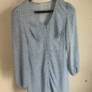 Blåvit mönstrad klänning från Nelly x Galmorous  - Supersöt blå kortklänning med vitt prickigt mönster. Klänningen har v-ringning, långa ärmar med lätt puff och markerad midjan, knäppning i fram. Perfekt för vår och sommar. Strl XS/34. 